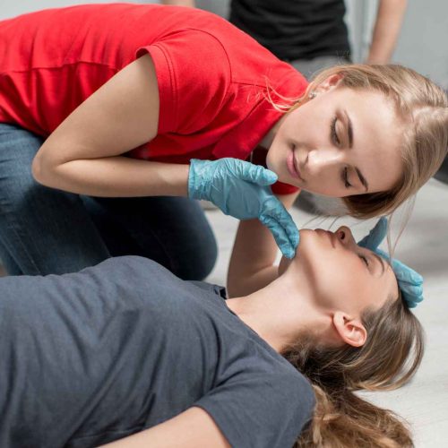 woman in red shirt wearing blue nitrile gloves performing a head-tilt-chin-lift on another woman laying on the ground. the woman in red is listening for the other woman's breathing and checking to see if her chest is rising and falling as part of CPR's ABCs. Coast2Coast