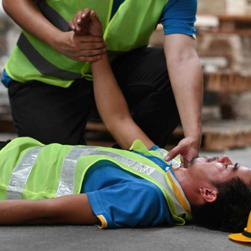 Workers in high-visibility vests providing first aid to an injured colleague at a warehouse, showing why first aid training is important
