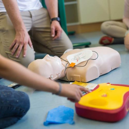People are gathered around a CPR training mannequin and a defibrillator. One person points at the defibrillator, while others observe. The setting appears to be a training session, with the focus on emergency response techniques.