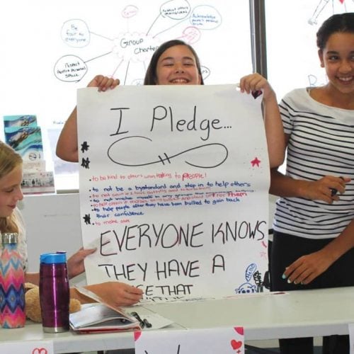 Three girls in a classroom setting, one seated with a notebook, and two standing, holding a large poster that reads "I Pledge... Everyone knows they have a voice..." The background shows a window and a whiteboard with doodles.
