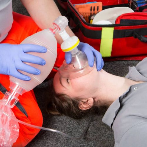 A person in a gray shirt receiving CPR, with another person wearing orange pants and blue gloves using a bag valve mask to administer breaths. A medical kit is open on the ground nearby.