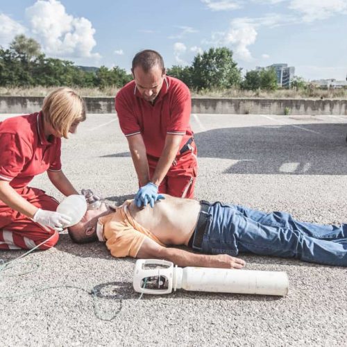 Two emergency responders in red uniforms perform CPR on a man lying on the ground. One administers chest compressions while the other uses a bag valve mask for ventilation. An ambulance is partially visible nearby.
