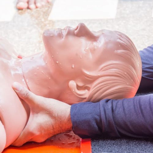 CPR training on a dummy, with two people's hands positioning the head for airway management. The dummy is on a red board, and the scene is well-lit, focusing on the importance of life-saving techniques.