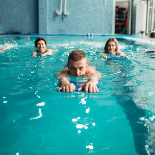 A group of people swimming in a pool, using kickboards. They are aligned in a row, each focusing on swimming. The pool area has blue tiles and a modern look, with some equipment visible in the background.