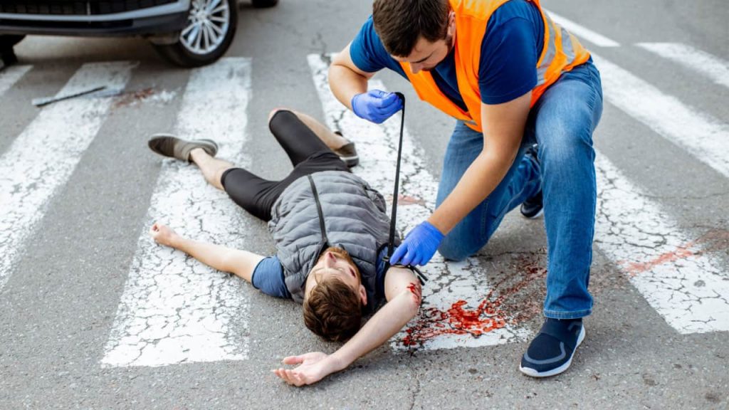 A person in a safety vest and blue gloves kneels on a crosswalk, tending to an injured person lying on the ground. Blood is visible on the pavement. The injured person is wearing a gray vest and dark pants. A vehicle is nearby.