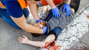 Two individuals in orange safety vests and blue gloves are assisting an injured person lying on a cracked, blood-stained pavement. One holds the person's head, while the other checks their torso. The person’s arm is outstretched.