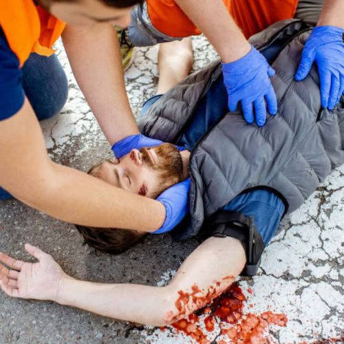 Two individuals in orange safety vests and blue gloves are assisting an injured person lying on a cracked, blood-stained pavement. One holds the person's head, while the other checks their torso. The person’s arm is outstretched.