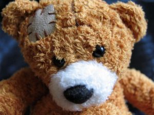Close-up of a well-loved teddy bear with a patch sewn on its head. The bear has soft, brown fur, shiny black eyes, and a white snout, creating a cute and endearing appearance. The background is a blurred dark blue color. Highlighting a head injury - Picture by Coast2Coast First Aid