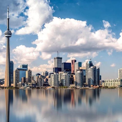 Toronto skyline with the CN Tower and various skyscrapers under a blue sky with clouds. Buildings are reflected in the calm water in the foreground, creating a mirrored effect.