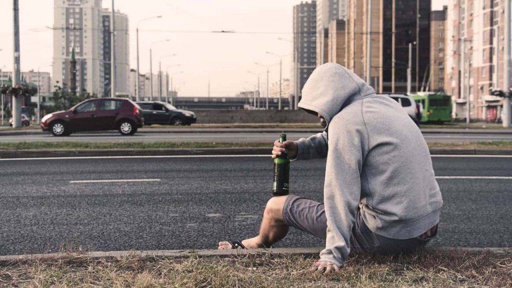 A person wearing a gray hoodie sits on a grassy area beside a city street, holding a green bottle. Cars are passing by, and tall buildings are visible in the background. The scene suggests an urban environment.