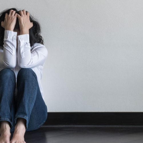 A person sits on the floor against a plain wall, dressed in a white shirt and blue jeans. They have their knees up and head in hands, conveying a sense of distress or overwhelming emotion. Barefoot, they appear deep in thought or anxiety.