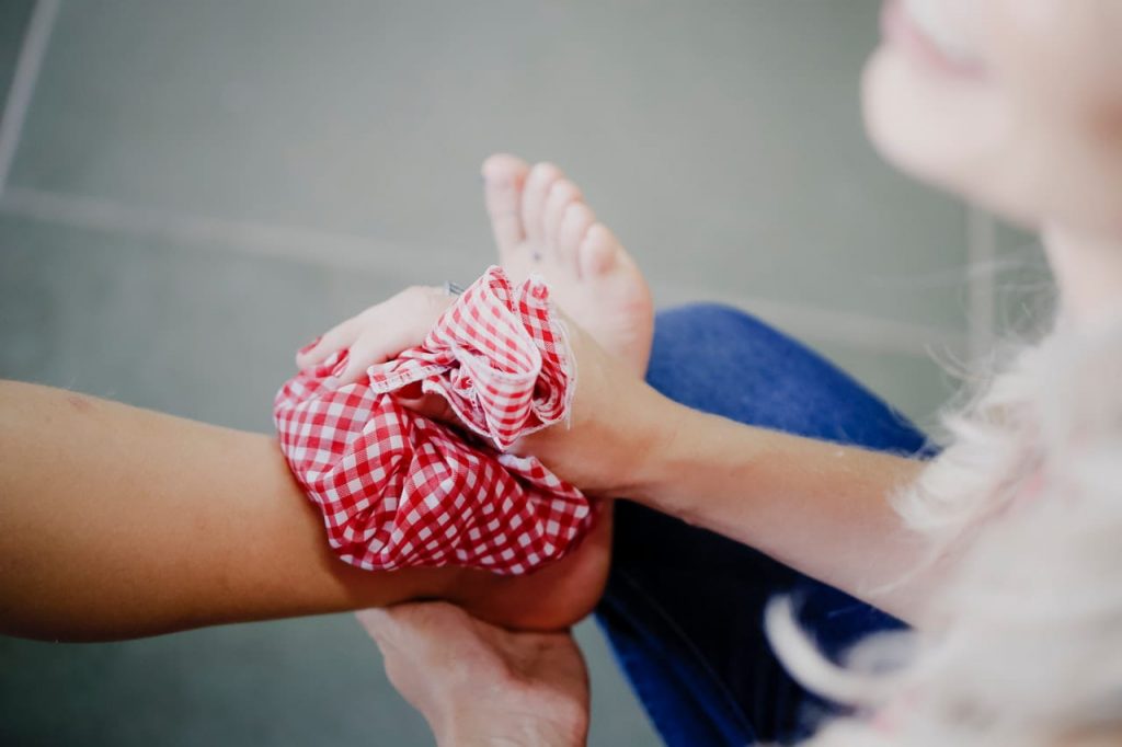A person holding a red and white checkered cloth wrapped around their ankle. The person is wearing jeans, and the background is a tiled floor. The focus is on the ankle and cloth, with the face slightly out of frame.