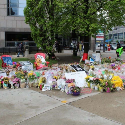 A large memorial on a city sidewalk features numerous flowers, candles, stuffed toys, and heartfelt messages. People walk nearby, and buildings are visible in the background.