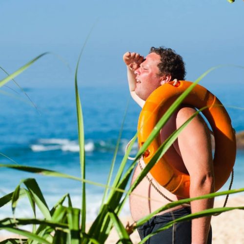 A man wearing an orange lifebuoy stands on a sandy beach, shielding his eyes from the sun. He faces the ocean, surrounded by greenery. The sea is calm, and the sky is clear and bright.
