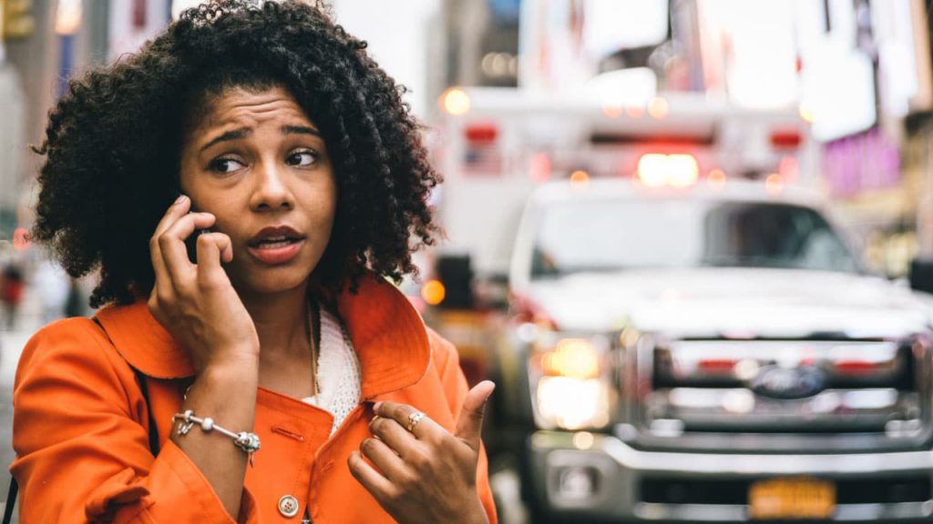 A woman with curly hair, wearing an orange coat, is talking on her phone. She looks concerned and is gesturing with her hand. An ambulance with flashing lights is visible in the background on a city street.