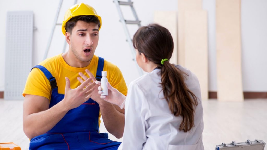 A construction worker wearing a yellow hard hat and blue overalls is sitting on the floor, gesturing animatedly. A person in a white coat is facing him, holding a small white bottle. A ladder and wooden boards are in the background.