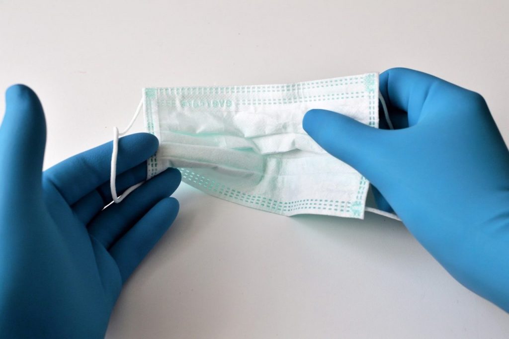 Two gloved hands holding a light blue surgical mask against a white background. The gloved hands are positioned to put on or adjust the mask, which has ear loops on each side.