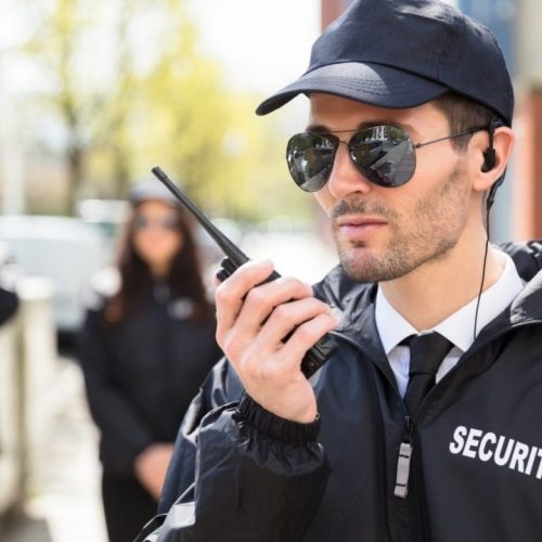 A man wearing sunglasses and a cap, dressed in a security jacket, talks into a walkie-talkie. In the background, three other security personnel stand out of focus. They all appear to be outdoors, near a building.