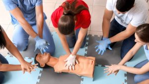 A group of people practicing CPR on a training mannequin. They are positioned around the mannequin on a foam mat, with one person performing chest compressions. Others are observing and preparing to practice. All are wearing casual clothes.