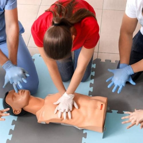 A group of people practicing CPR on a training mannequin. They are positioned around the mannequin on a foam mat, with one person performing chest compressions. Others are observing and preparing to practice. All are wearing casual clothes.