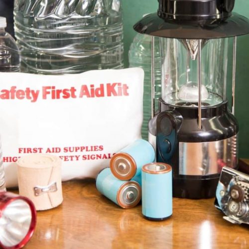Emergency preparedness supplies including bottled water, a "Safety First Aid Kit," batteries, a red flashlight, a lantern, canned goods, a can opener, and a radio are arranged on a table.