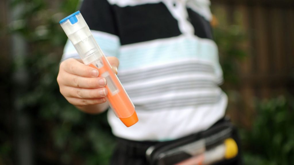 A person holding a cylindrical orange and clear device, with a focus on the hand and device. The person is wearing a striped shirt and has a black belt bag visible in the lower part of the image. The background is blurred greenery.