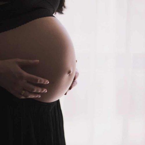 A close-up side view of a pregnant person gently holding their bare belly with both hands. They are wearing a black top and skirt. Soft light filters through a sheer curtain in the background.