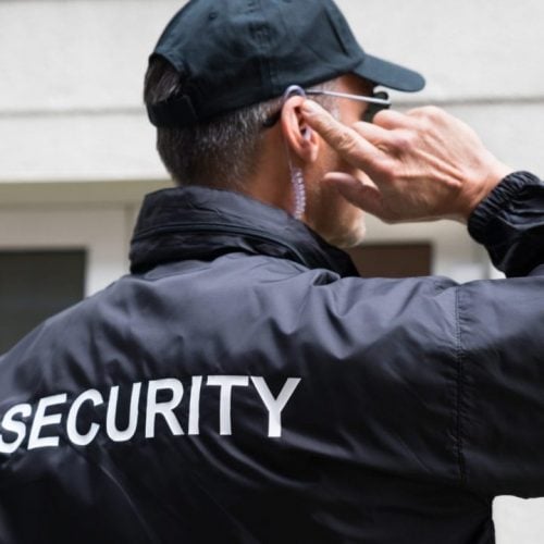 A security guard wearing a black jacket with "SECURITY" written on the back and a black cap stands outside a building. They are holding an earpiece with their right hand, appearing attentive and focused.