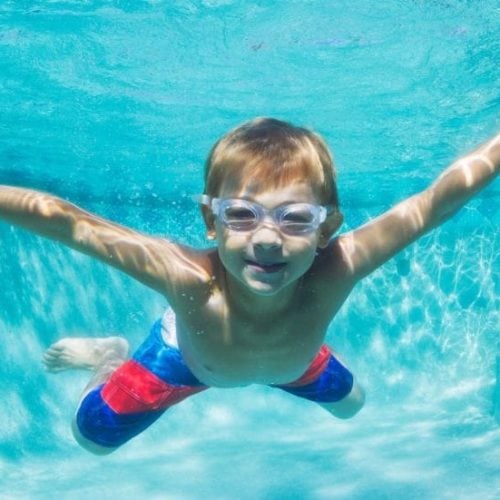 A young boy swims underwater in a pool, wearing goggles and red and blue swim trunks. He is smiling, with his arms outstretched, surrounded by clear blue water.