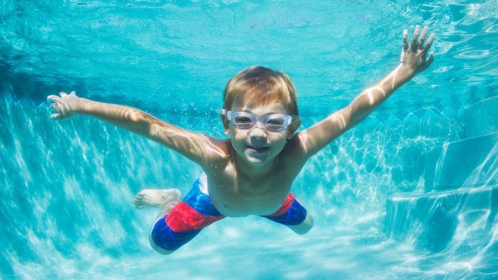 A young boy swims underwater in a pool, wearing goggles and red and blue swim trunks. He is smiling, with his arms outstretched, surrounded by clear blue water.