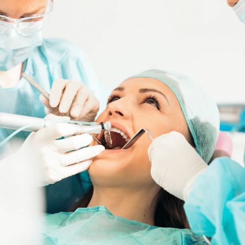 A woman is receiving dental treatment from two dentists. She is lying back with her mouth open as they work, wearing blue surgical gowns, gloves, and masks. The setting appears to be a dental clinic.