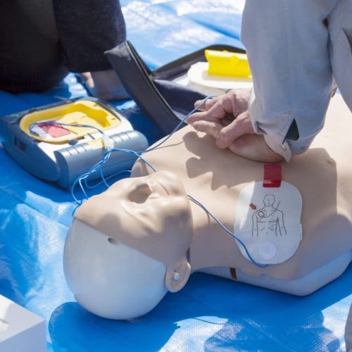 Person demonstrating CPR on a mannequin with an AED device next to them, placed on a blue tarp. The mannequin has electrode pads on its chest, and the scene indicates a training session.