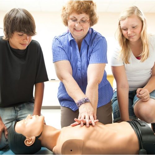 A woman demonstrates CPR on a mannequin while two teenagers watch attentively. The group is in a classroom setting with some furniture and a whiteboard visible in the background.