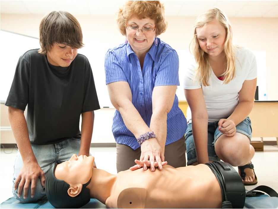 A woman demonstrates CPR on a mannequin while two teenagers watch attentively. The group is in a classroom setting with some furniture and a whiteboard visible in the background.