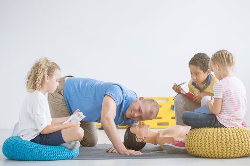 A man demonstrates CPR on a dummy to three children. They sit on cushions and observe, with two holding notebooks. The setting is bright, with a yellow stretcher visible in the background.