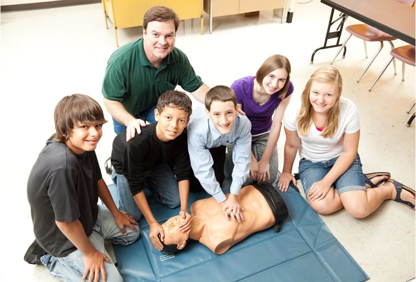 A group of students and an adult instructor are gathered around a CPR training manikin. The students are practicing CPR techniques, with some placing their hands on the manikin's chest. They are indoors on a tiled floor.