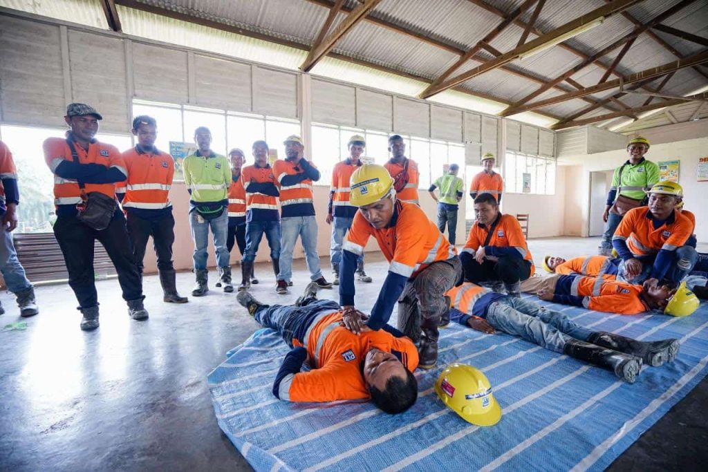 Construction workers in orange safety gear participate in a first aid training session. One worker demonstrates a technique on a colleague lying on a blue mat, while others observe. The setting is an indoor area with large windows and a corrugated metal roof.