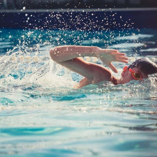 A person wearing goggles swims freestyle in an outdoor pool, creating splashes of water. The sunlight reflects off the water surface, and lane dividers are visible in the background.