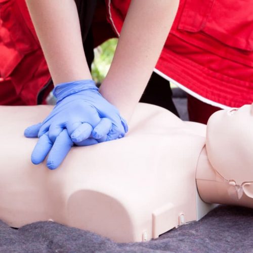 Person in red clothing and blue gloves performing CPR on a practice dummy outdoors. The dummy is lying on a dark surface, and the person is applying chest compressions with interlocked hands.
