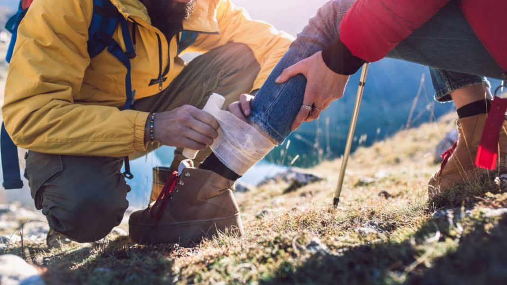 Person in a yellow jacket bandages the ankle of another person sitting on the grass, who is wearing jeans and hiking boots. They are outdoors with trekking poles, and the background shows hills under a sunny sky.