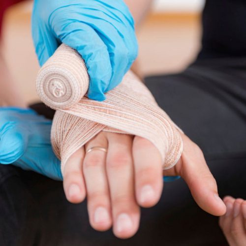 A person with gloved hands is wrapping a white bandage around another person's injured hand. The seated person has their hands resting on their lap, and the background is out of focus.