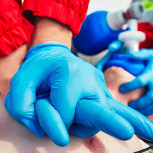 A person wearing blue gloves performs CPR chest compressions on another individual lying on the ground. A second person assists with a bag-valve mask in the background, providing ventilation. Both wear red uniforms, indicating emergency medical services.