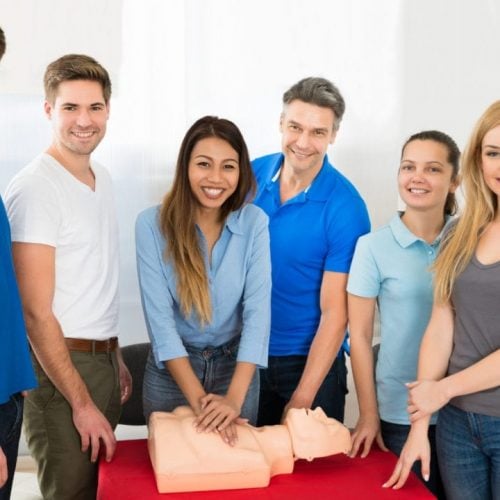 A group of six people smiles near a CPR mannequin on a table. One person demonstrates CPR by pressing on the mannequin's chest. They are in a bright room, with casual attire. A clock is visible on the wall.