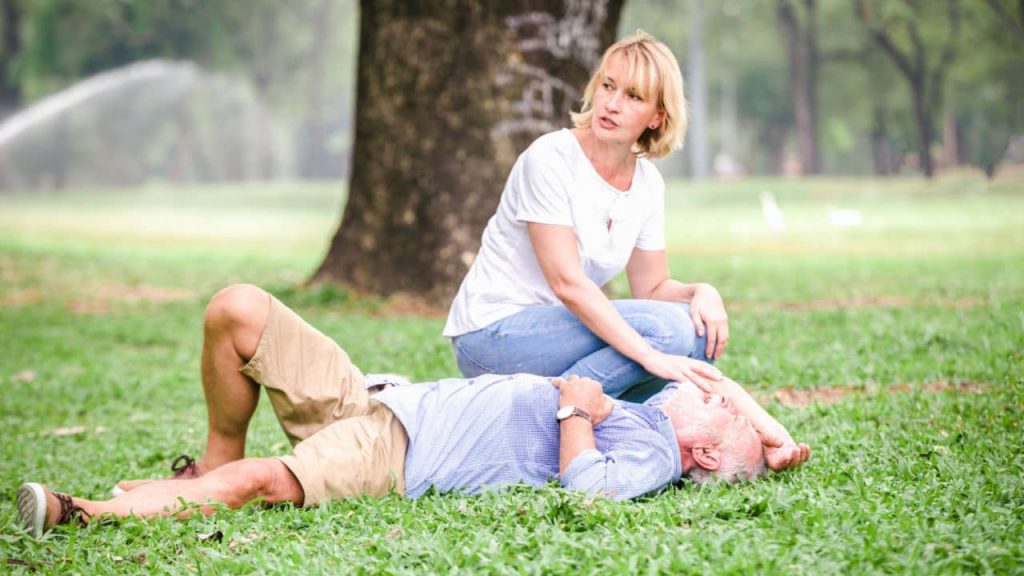 A woman kneels on the grass in a park, offering assistance to an elderly man lying on his back with his eyes closed. She has her hand on his forehead, appearing concerned. A large tree trunk is in the background, with park greenery and a water fountain visible. 