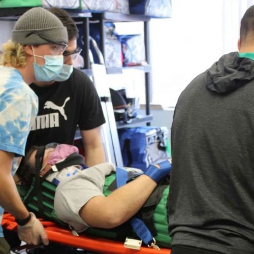 A group of people, wearing masks and gloves, are helping a person on a stretcher. The person on the stretcher is secured with straps, and the setting appears to be indoors, possibly in a medical or training facility.