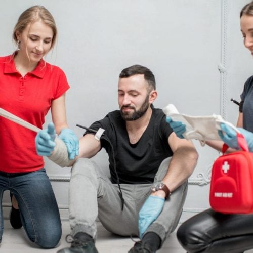 Two women in casual attire and gloves assist a seated man with a bandaged arm. One woman is holding the bandage, and the other has a first aid kit. They are indoors against a light-colored wall.