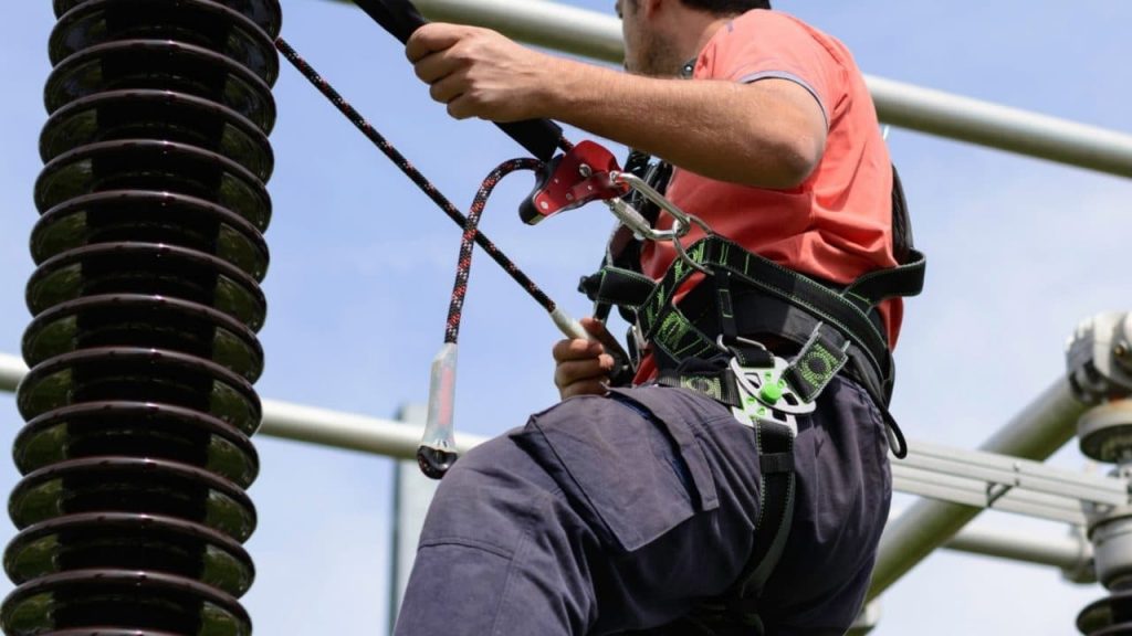 A person wearing a safety harness climbs a large structure, secured by ropes. They are looking upwards, dressed in a red shirt and dark pants, against a clear blue sky.