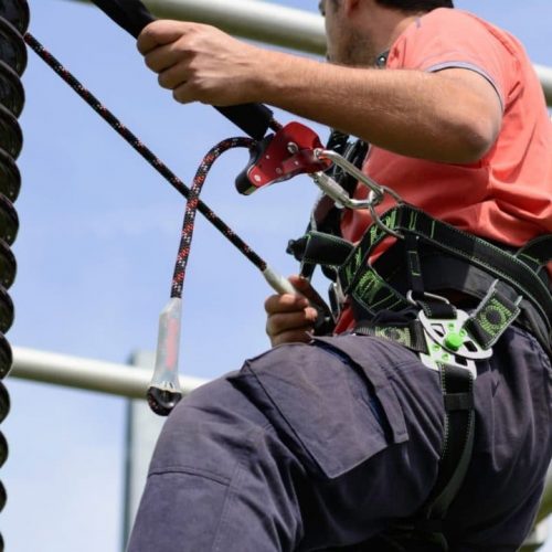 A person wearing a safety harness climbs a large structure, secured by ropes. They are looking upwards, dressed in a red shirt and dark pants, against a clear blue sky.