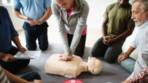 A diverse group of people kneels around a CPR manikin. A woman in the center performs chest compressions. A man holds a CPR instruction sheet. A red medical kit with a white cross is on the floor nearby.