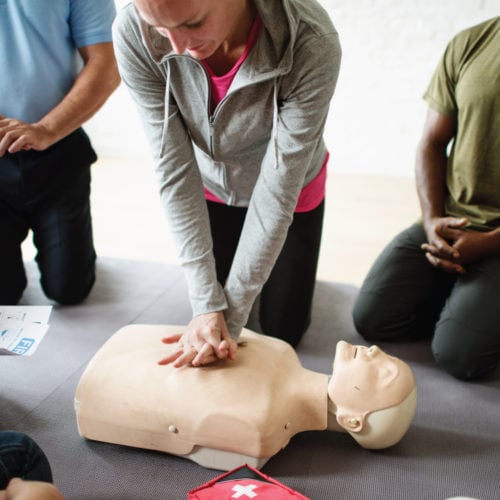 A diverse group of people kneels around a CPR manikin. A woman in the center performs chest compressions. A man holds a CPR instruction sheet. A red medical kit with a white cross is on the floor nearby.